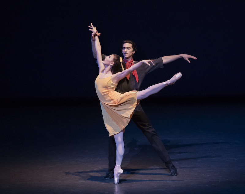 Tiler Peck in a yellow dress in a high arabesque en pointe. Zachary Catazaro holds her wrist and wears a red scarf around his neck.